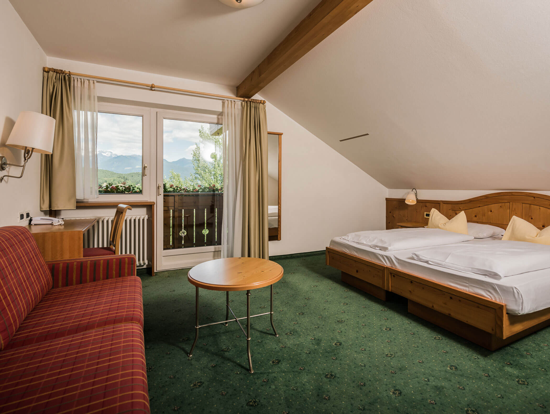 On the right a red sofa, in front of it a small round table and on the left a double bed, in the background a balcony door - Wirtshaushotel Alpenrose