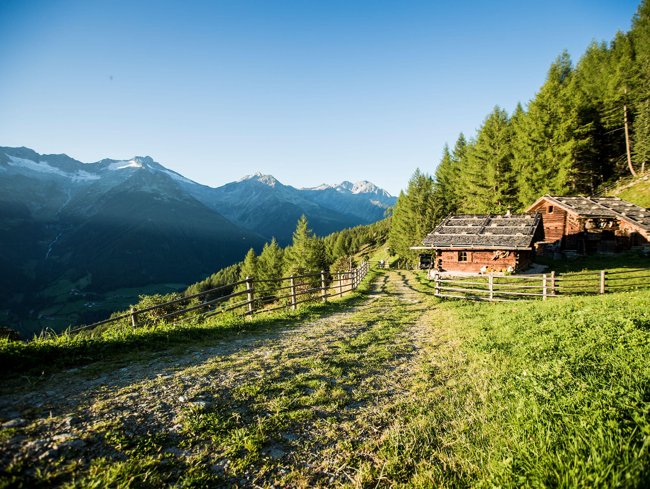 Hiking trail in the Dolomites with two wooden huts and forest on the right and mountains in the background - Wirtshaushotel Alpenrose
