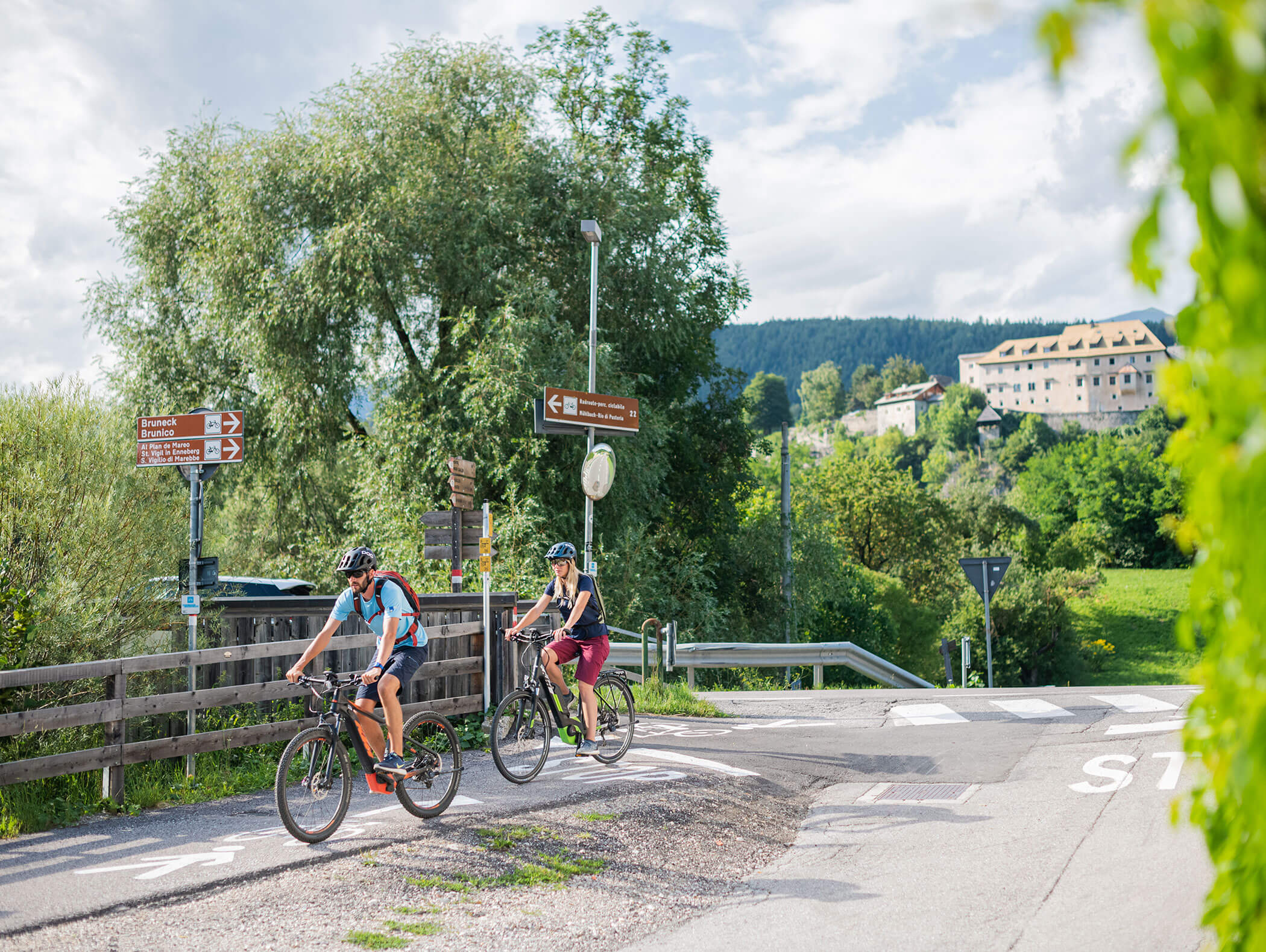 Cycle path in the Val Pusteria with two cyclists in the foreground and a building in the background - Wirtshaushotel Alpenrose