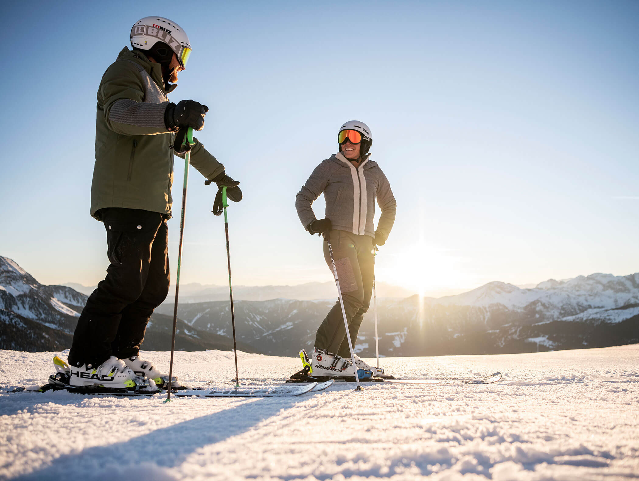 Two skiers on the slopes of Kronplatz with snow-covered mountains and the sun in the background - Wirtshaushotel Alpenrose
