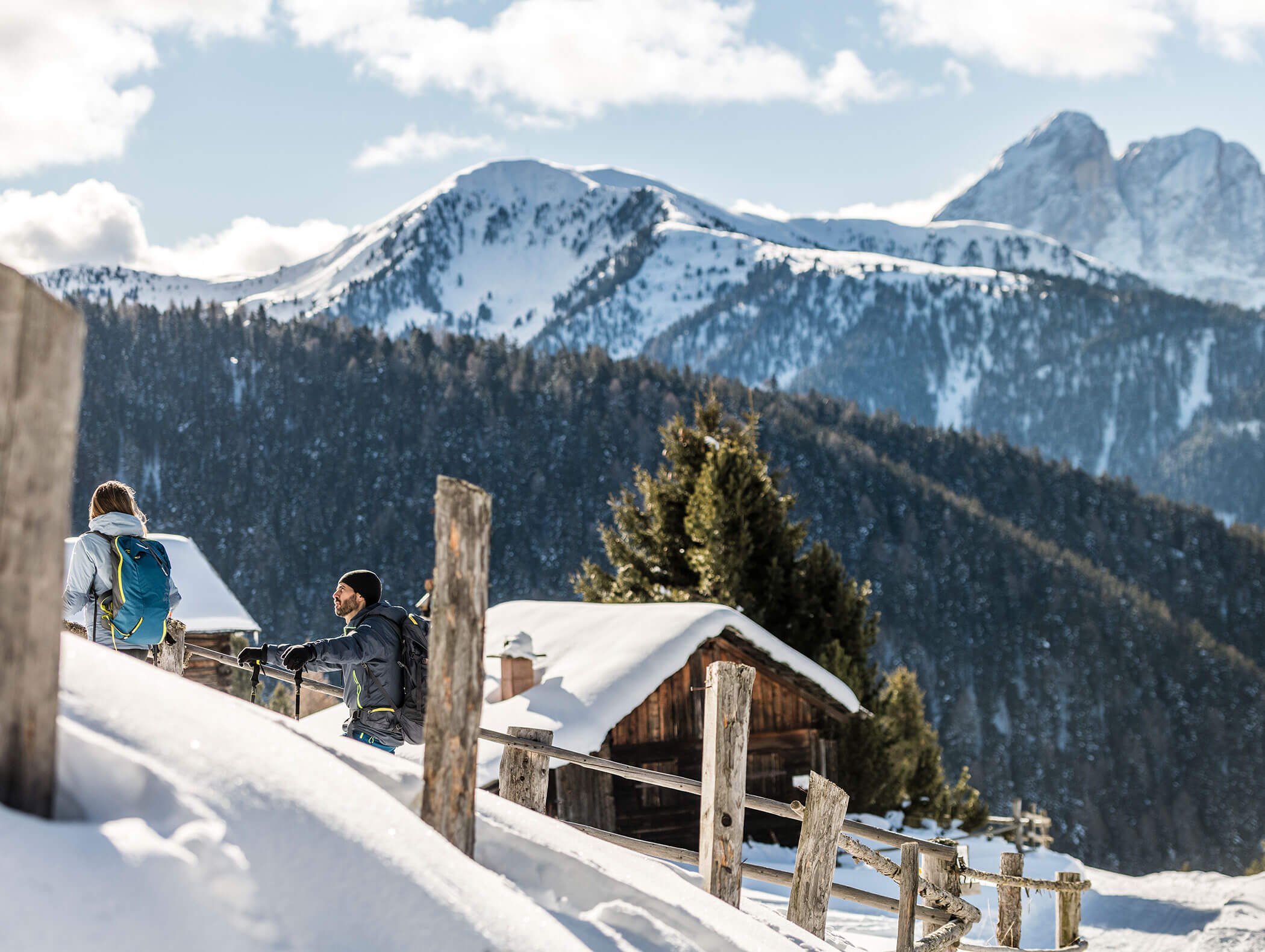 Two people in winter with a wooden fence in the foreground and a wooden hut, mountains and forests in the background - Wirtshaushotel Alpenrose